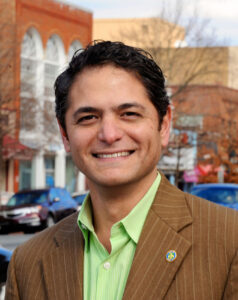 A smiling man in a green shirt and brown suit jacket.